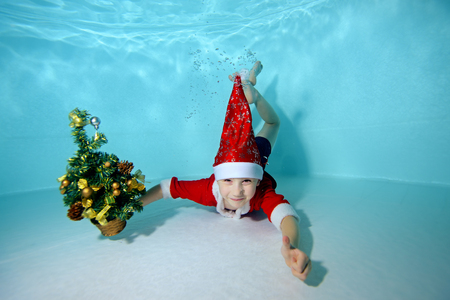Happy little boy lying underwater at the bottom of the pool, holding Christmas tree in hand and smiling. Portrait. Landscape orientation.の写真素材