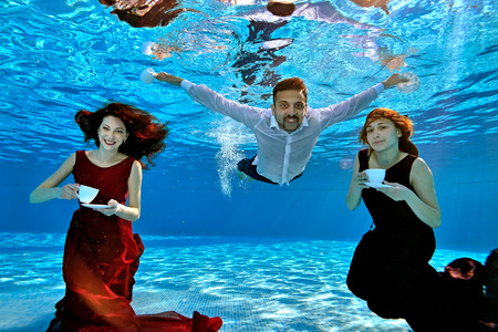 Two beautiful girls in a red and Burgundy dress and a guy in a white shirt swim and play underwater in the pool. They hold white cups and saucers in their hands, look at me and smile. Portrait.の写真素材