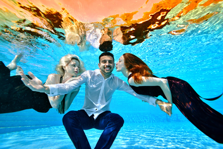 Two young girls in love, a blonde and a brunette, swim in dresses and play with a guy in a white shirt under water in the pool. They hug and kiss him. The guy looks at the camera and smiles. Portrait.の写真素材