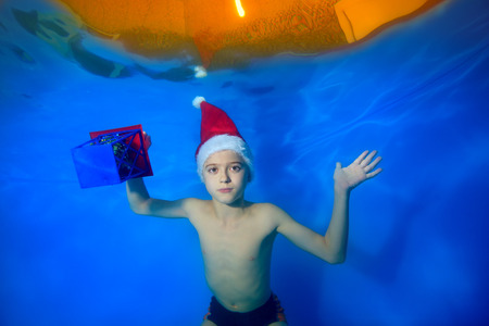 A little boy swims under the water in the pool in a red Christmas cap and with a gift in his hand on a blue background and looks at the camera. Portrait. Concept. Landscape orientation of the image.の写真素材