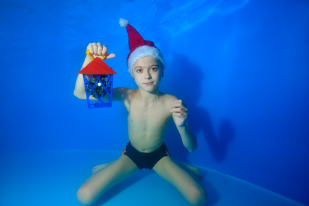 Portrait of a little boy in a red Christmas cap with a gift in his hand on a blue background. He swims and poses underwater at the bottom of the pool and looks into the camera. Concept.の写真素材