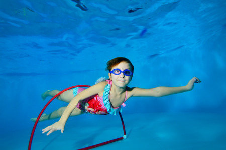 The child goes in for sports and swims underwater through a red Hoop at the bottom of the pool on a blue background and looks at me. Portrait. Landscape orientation. Underwater photography.の写真素材