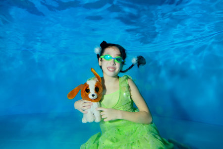 A little girl swims and poses underwater in a green dress and swimming glasses at the bottom of the pool, holds a toy - dog in her hands, looks at the camera and smiles. Portrait. Shooting underwater.の写真素材