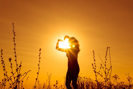 Silhouette against the sunlight of a girl who stands at sunset in a field among the grass and holds her hands over her head in the shape of a heart. Horizontal orientation of the image.の写真素材