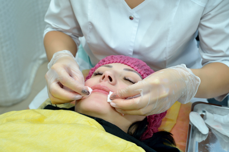 A young woman lies in a beauty salon, and a cosmetologist wipes her lips and skin with cotton swabs after a permanent lip makeup procedure. Portrait. Close up. Horizontal view.の写真素材