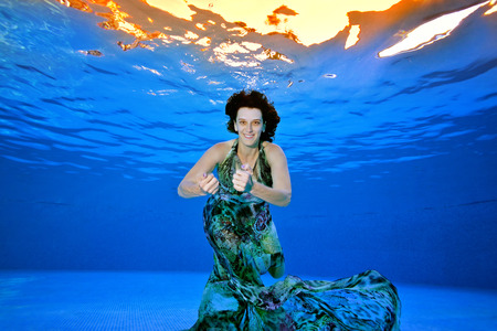 Cute girl in a fashionable dress posing underwater in the pool on a blue background. She looks at the camera and smiles. Portrait. Shooting underwater.の写真素材