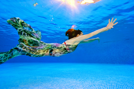 Girl model swims and posing underwater in the pool in a dress on a blue background. Shooting underwater. Side view. Horizontal orientation of the photo.の写真素材