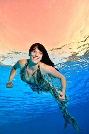 A woman in a beautiful dress swims underwater in the pool against the sunlight on the surface, looking at the camera and smiling. Portrait. Shooting underwater. Vertical orientation.の写真素材