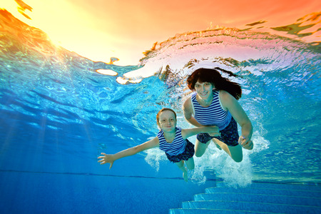 Happy family, mother and daughter, play sports on vacation and swim underwater in the pool in striped swimsuits on a bright Sunny day. They look at the camera and smile. Portrait. Bottom view.の写真素材