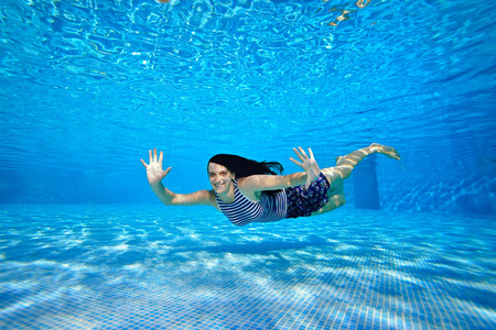 A cheerful sports girl swims under the water near the bottom of the pool in a striped swimsuit on a bright Sunny day, looks at the camera, waves her hands and smiles. Portrait. Underwater photography.の写真素材