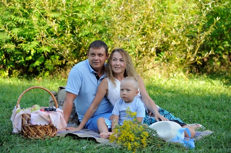 Dad, mom and little boy are sitting on the grass in the Park on a summer day, looking away and smiling. Family picnic in nature. Portrait. Horizontal view.の写真素材