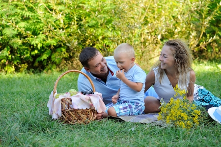 A little boy eats grapes, and mom and dad sit and look at him in the Park on a summer day and smile. Family picnic in nature. Portrait. Horizontal view.の写真素材