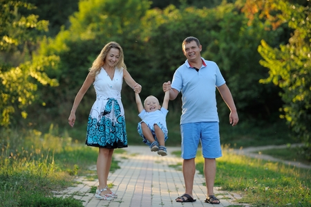 Mom, dad and baby are walking in the Park at sunset on a summer day. Parents shake the child in his arms, as on a swing. Concept. Close up. Horizontal view.の写真素材