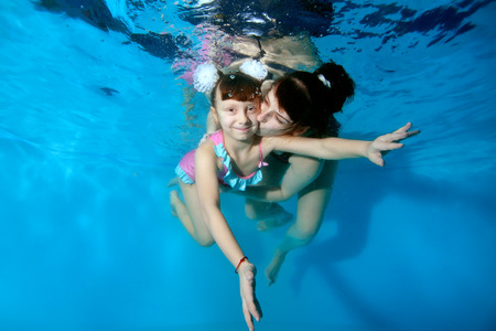 Happy mother kissing and hugging her young daughter under water in the pool. They play sports and smile on a blue background with their eyes open. Portrait. Horizontal view.の写真素材