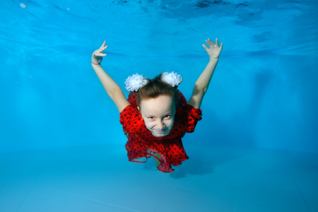 Smiling little girl swims and dives underwater in the pool with her eyes open in red dress on blue background. Portrait. Horizontal view.の写真素材