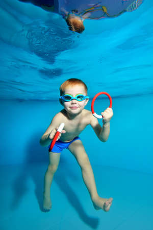 A little boy goes in for sports and swims underwater in the pool near the water surface. Portrait. Underwater photography. Blue background. Vertical orientation.の写真素材