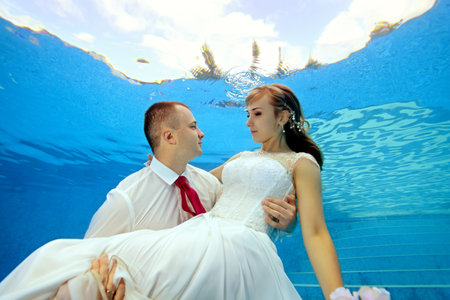 Lovers bride and groom in the pool underwater on a Sunny day. The groom holds the bride in his arms, the young look at each other. Portrait. Concept. Wedding underwater. Horizontal view.の写真素材