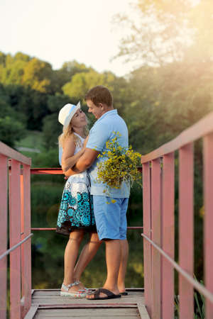 A woman and a man in love look at each other tenderly in the Park by the water at the sunset of a summer day. Bouquet of flowers in the hands of the girl. Photo in full growth. Portrait. Vertical viewの写真素材