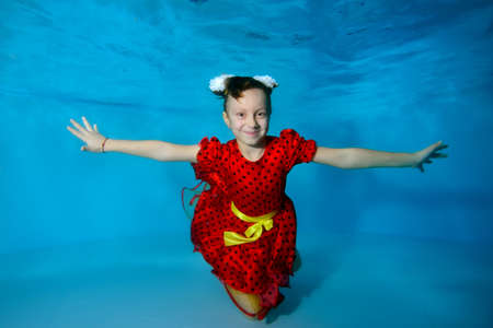 A cute little girl is swimming underwater at the bottom of the pool with her eyes open in a red dress. She smiles and poses for the camera. Hand in hand. Portrait. Horizontal view.の写真素材