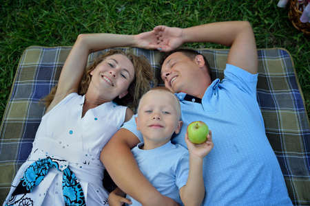 Happy family lying and dreaming on the grass in the Park at sunset summer day. Parents hold hands and smile. The child holds an Apple. Portrait. The view from the top. Horizontal view.の写真素材