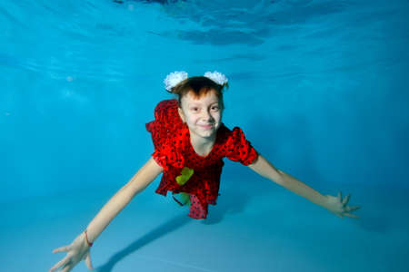 Laughing girl swims underwater at the bottom of the pool with her eyes open in a red dress, with bows on her head. She's posing for the camera. Portrait. Horizontal view.の写真素材