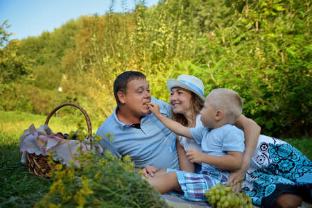 Happy family picnic in the park on summer day. Mom, dad and a little boy. The boy feeds the Pope grapes and laughing along with them. Family picnic in nature. Portrait. Horizontal view.の写真素材
