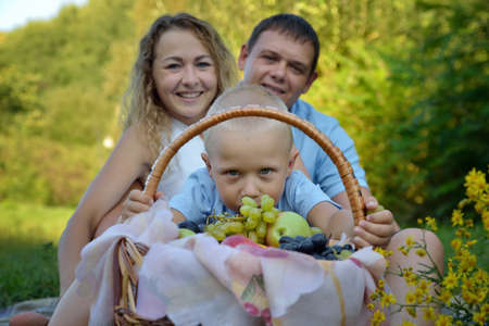 A little boy looks through a fruit basket, and mom and dad sit behind him on the grass in the Park, smiling and looking at the camera at sunset. Family picnic in nature. Portrait. Horizontal view.の写真素材