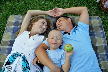 Mom, dad and little son lie and dream on the grass in the Park at sunset summer day. Parents look at each other and smile. The child holds an Apple. Portrait. The view from the top. Horizontal view.の写真素材