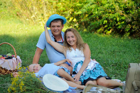 Happy family having fun playing sitting in the Park on a summer day. Mom puts on dad's women's hat and laughs with his son. Family picnic in nature. Portrait. Horizontal view.の写真素材