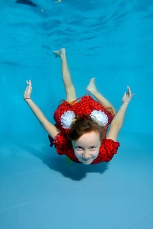 A little girl diving to the bottom of the pool in a red dress, with bows on her head, her eyes open. She's posing for the camera. Portrait. Vertical view.の写真素材