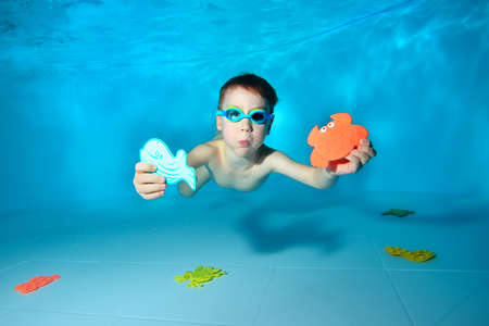 Cute sports baby swims underwater and holds sea toys in his hand. He looks at the camera and smiles. Portrait. Underwater photography. Horizontal orientation.の写真素材
