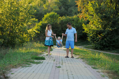 Parents swing the little boy in their arms like a swing and walk along the path in the Park at the sunset of a summer day. The concept of family happiness. Shut. Horizontal view.の写真素材