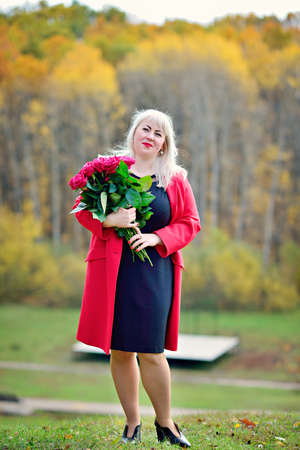 Beautiful woman, blonde in a red coat with a bouquet of red roses in the Park. Sunny day. Size plus xxl female. Close up. Vertical view.の写真素材