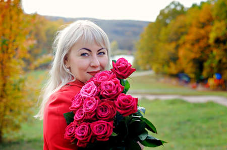 A pretty woman, blonde, stands in a Park in a red coat, holding a bouquet of red roses to her face. Sunny day. Size plus xxl female. Close up. Horizontal view.の写真素材