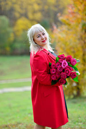 Cute middle-aged girl, blonde in the Park in a red coat with a bouquet of red roses on the background of autumn trees. Stands sideways and looks at the camera. Size plus xxl female. Vertical view.の写真素材