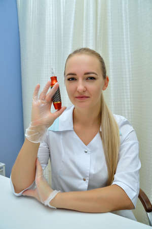 Beautiful girl beautician in a white medical gown, sitting at a white table, on a white background, holding a cosmetic tool in his hand and looking at the camera. Portrait. Vertical view.の写真素材