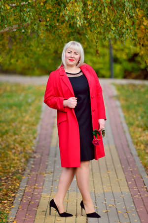 A pretty big blonde in a black dress and a red coat with a rose in her hands is standing on the path in the Park. Woman's size plus xxl. General plan. Vertical view.の写真素材