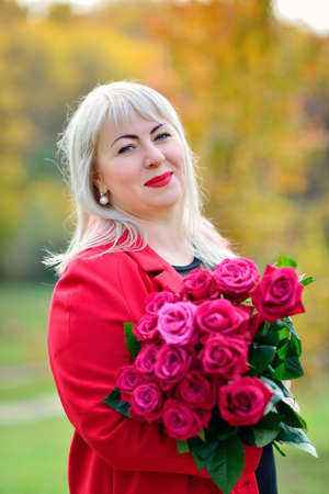 Portrait of a pretty woman, blonde in a red coat with a bouquet of red roses in the Park. Sunny day. Size plus xxl female. Close up. Vertical view.の写真素材