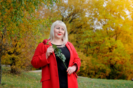 A large beautiful blonde in a red coat and a scarlet rose in her hand, in the Park on a Sunny autumn day looks dreamily to the side. Woman's size plus xxl. Horizontal view.の写真素材