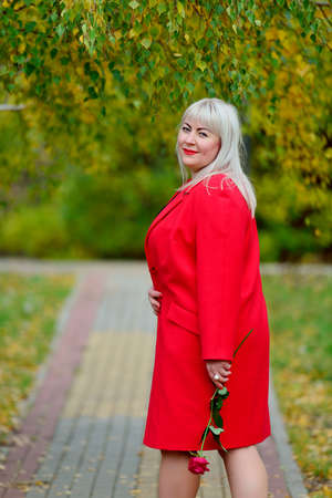 A cute blonde in a red coat and a scarlet rose in her hand, in the Park on a Sunny autumn day dreamily looks at the frame. Woman's size plus xxl. Horizontal view.の写真素材
