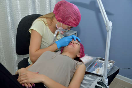Master beautician, blonde, performs the procedure of microblading eyebrows girl in a beauty salon with a cosmetic tool. Portrait. Close up. Horizontal view.の写真素材