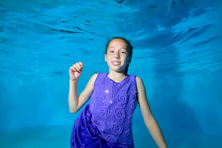 A little girl swims underwater in the pool. She smiles, looks at the camera with her eyes open. She's wearing a beautiful dress. Blue background. Portrait. Horizontal view.の写真素材