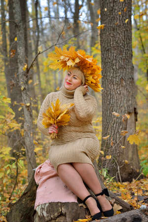 A sweet woman, a middle-aged blonde in autumn leaves, with a wreath on her head, sitting on a stump in the autumn forest in a gentle jumper. Size plus xxl female. Vertical view.の写真素材