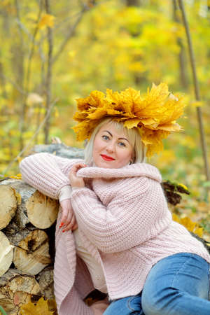 Portrait of a beautiful middle-aged blonde in a wreath of autumn leaves, who sits in a gentle jumper in the forest on a yellow background, looking at the camera and smiling. Woman's size plus xxl.の写真素材