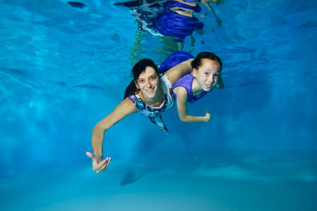 Mom and little daughter swim underwater in the pool. They hug, smile, look at the camera with their eyes open. They're wearing beautiful dresses. Portrait. Horizontal view.の写真素材