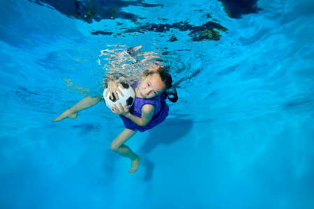 Cute girl swimming and playing underwater with the ball. On a blue background in a dress, eyes open. Portrait. Horizontal view.の写真素材