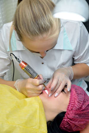 Woman-cosmetologist during the procedure of permanent makeup lips, which she makes European girl lying on the couch in the beauty salon.Concept. Vertical orientation of the image.の写真素材