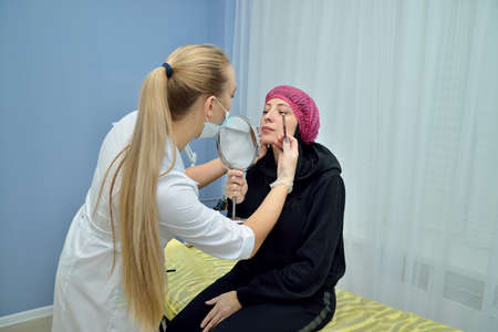 Beautician in a white coat prepares the patient for the procedure of permanent makeup lips in the beauty salon. She shows the outline drawn on her lips in the mirror. Portrait. Horizontal view.の写真素材