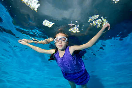Happy, smiling child. A girl in a purple dress and swimming goggles swims in the pool arms outstretched. Underwater photography. Horizontal view.の写真素材