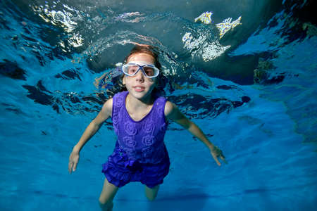 Cute little girl swimming underwater in the pool. She smiles and looks at the camera in a purple dress and swimming glasses. Blue background. Portrait. Horizontal view.の写真素材